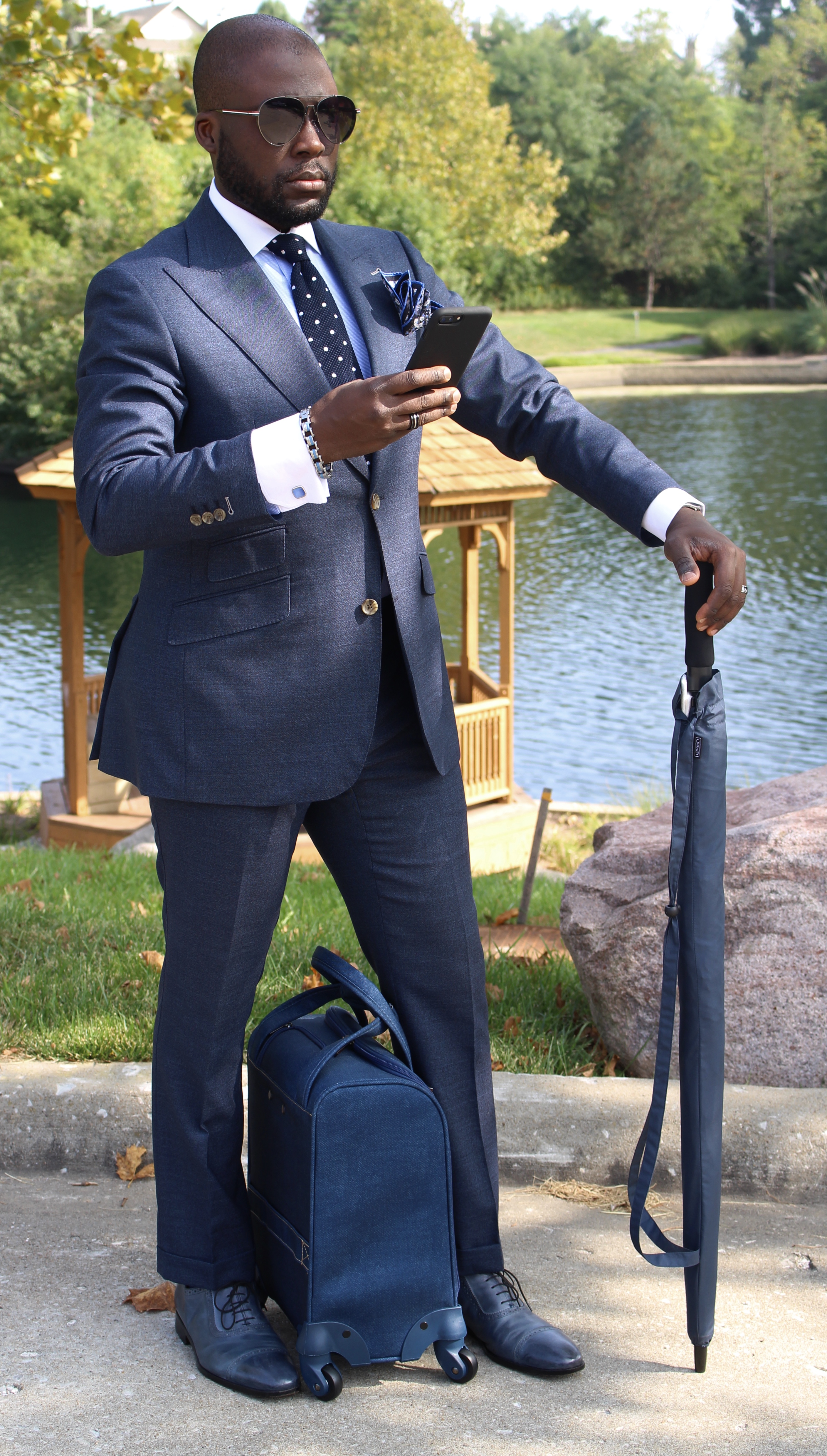 Photo 6 "The Navigator": dark blue suit, navy polka-dot tie, sunglasses, smartphone and umbrella, travel bag, outdoor setting by a pond with trees